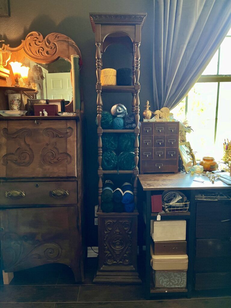 A 1970s wooden narrow shelf with intricate wood work is half full of yarn, standing between a desk with gold decorations and apothacery drawers, and an antique secretary desk with a mirror and flower lamp